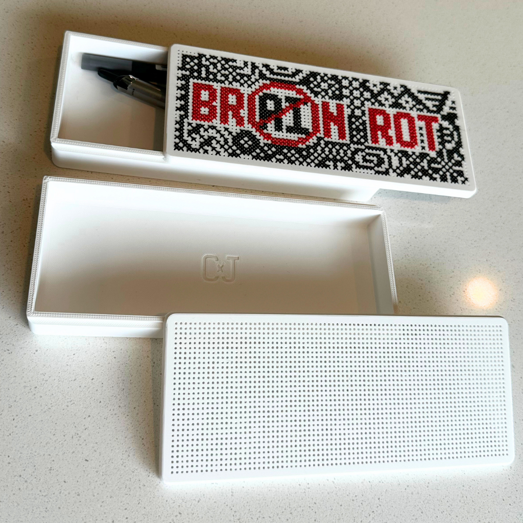 A white rectangular speaker and its matching open storage box are shown on a light speckled countertop. The box lid features a black-and-white pixelated design with the words 'BRAIN ROT' crossed out in red.