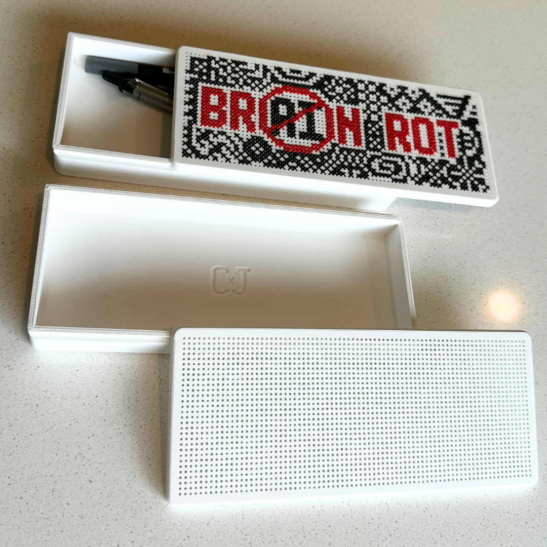 A white rectangular speaker and its matching open storage box are shown on a light speckled countertop. The box lid features a black-and-white pixelated design with the words 'BRAIN ROT' crossed out in red.
