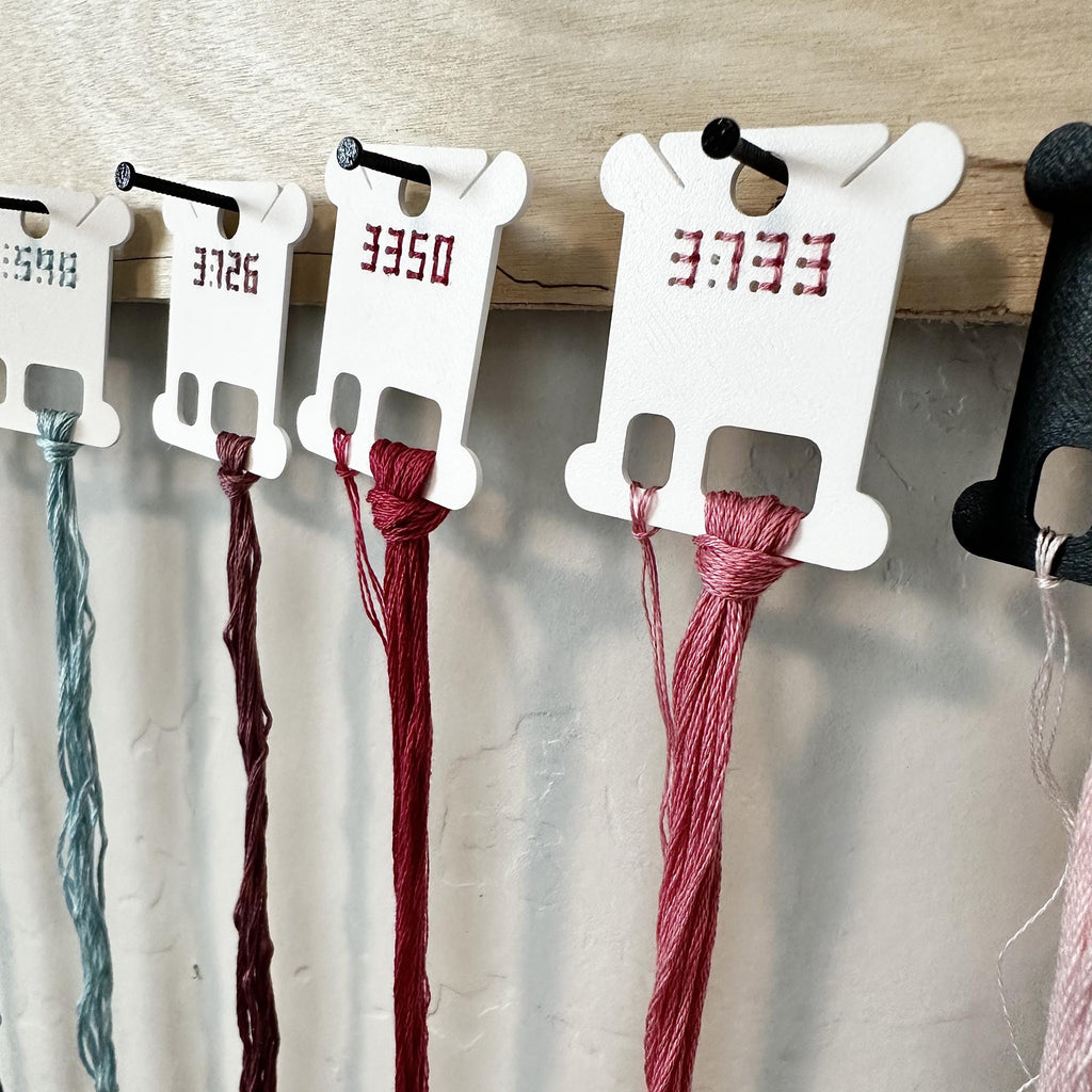 White plastic thread spool holders hanging on a wooden rack, each holding a different thread color including teal, burgundy, and dusty rose, with red printed numbers on the holders.