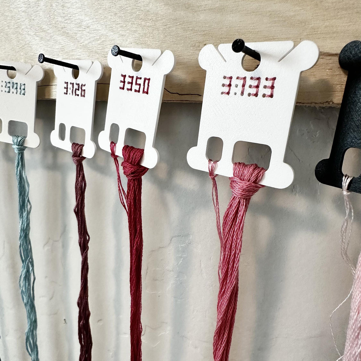 White plastic thread spool holders hanging on a wooden rack, each holding a different thread color including teal, burgundy, and dusty rose, with red printed numbers on the holders.