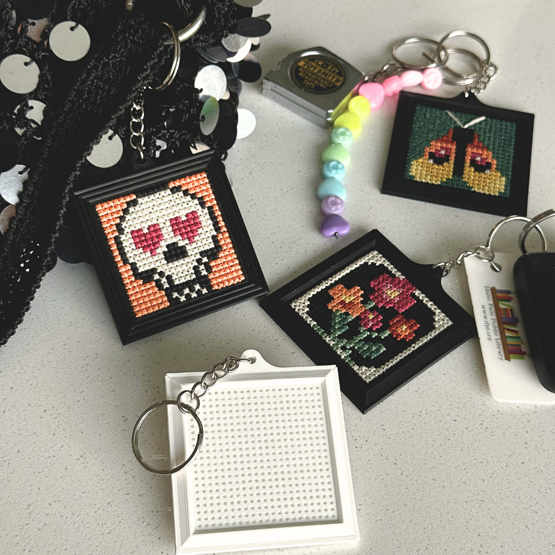 A collection of small cross-stitch keychains displayed on a light gray surface. The keychains include finished designs such as a skull with heart-shaped eyes, flowers, and a pair of crossed swords, alongside unfinished blank frames and a beaded charm.