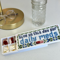 A white pill organizer case with a cross-stitch style label reading 'Give us this day our daily meds' in blue, green, and red, shown alongside a clear glass jar and eyeglasses on a light countertop.