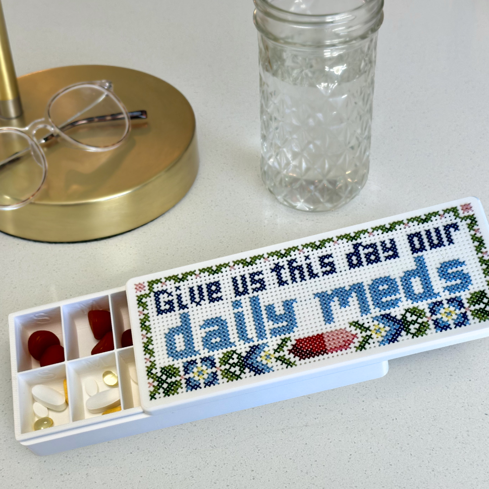 A white pill organizer case with a cross-stitch style label reading 'Give us this day our daily meds' in blue, green, and red, shown alongside a clear glass jar and eyeglasses on a light countertop.