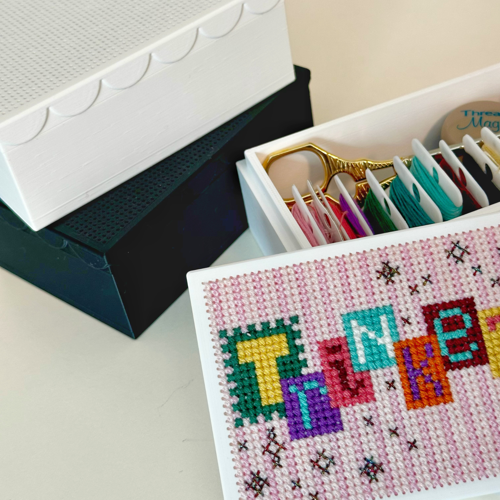 A white 3D printed craft organizer box with a scalloped lid, shown open with colorful embroidery floss inside and a decorated front panel featuring colorful letters spelling trinkets. 