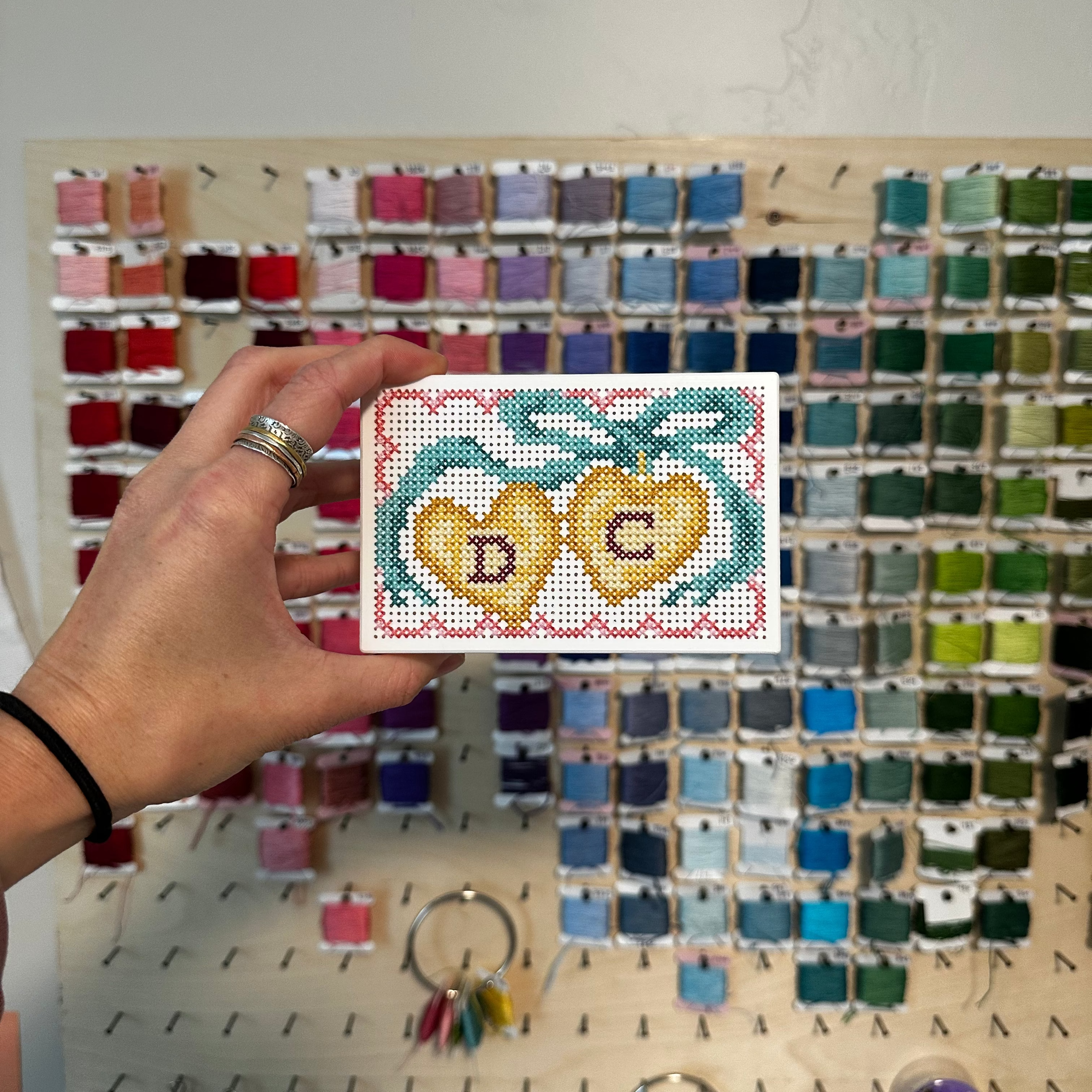 A hand holds a small cross-stitch sampler card featuring two yellow hearts with the initials 'D' and 'C' inside, set against a large wall display of many colorful embroidery floss bobbins. 