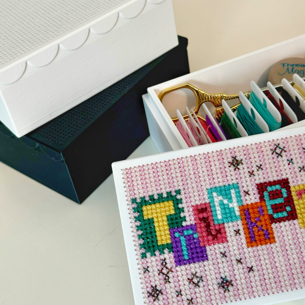A white 3D printed craft organizer box with a scalloped lid, shown open with colorful embroidery floss inside and a decorated front panel featuring colorful letters spelling trinkets.