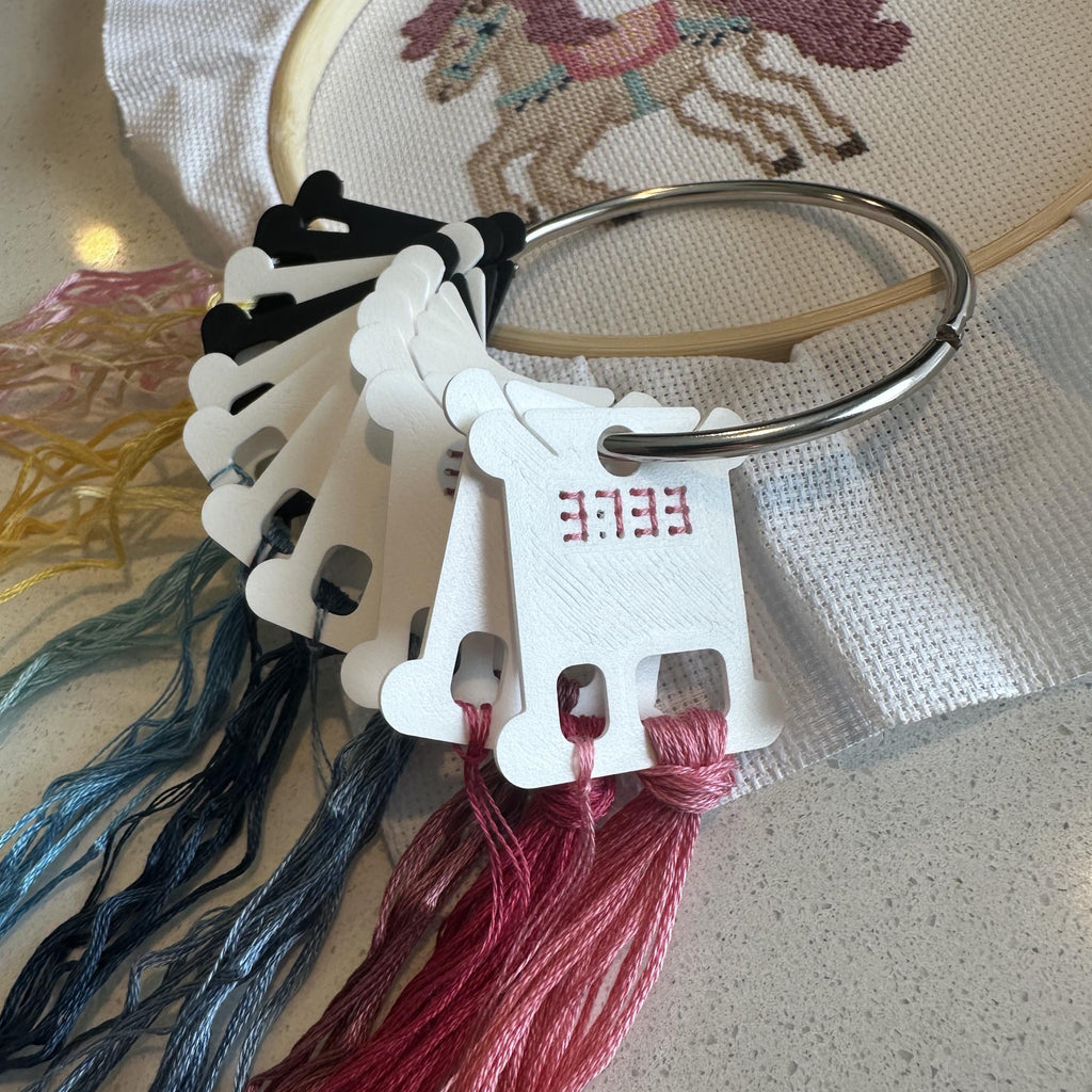 A close-up of embroidery floss skeins and a wooden embroidery hoop on a light surface. The hoop contains partially stitched cross-stitch artwork of a horse, and the floss tags show the letters EEEEE.