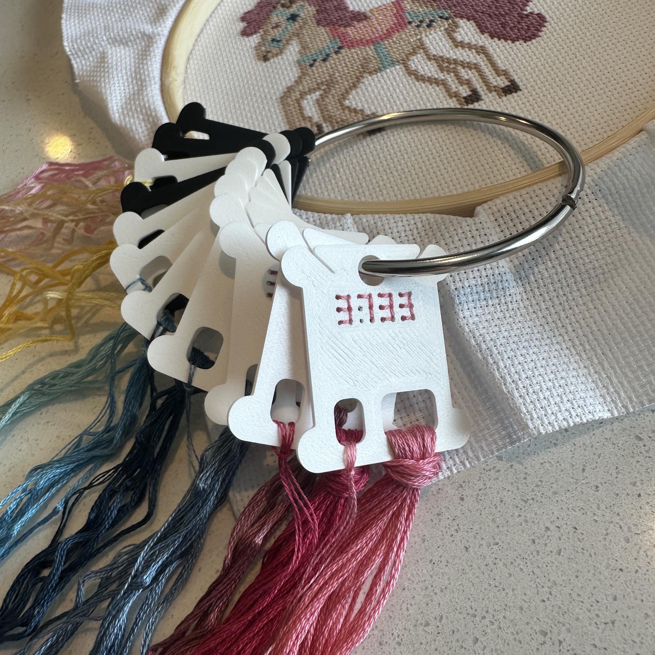 A close-up of embroidery floss skeins and a wooden embroidery hoop on a light surface. The hoop contains partially stitched cross-stitch artwork of a horse, and the floss tags show the letters EEEEE.