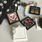 A collection of small cross-stitch keychains displayed on a light gray surface. The keychains include finished designs such as a skull with heart-shaped eyes, flowers, and a pair of crossed swords, alongside unfinished blank frames and a beaded charm.